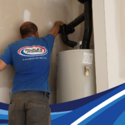 Plumber in a blue Poole’s Plumbing shirt repairs insulated pipes above a residential water heater.